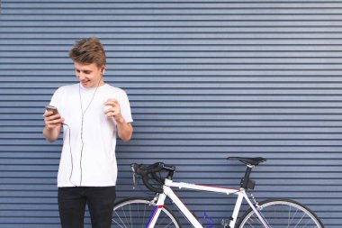 Mutlu genç öğrenci bir smartphone ve bir fincan kahve duvar arka planı elinde beyaz bisiklet yakınındaki ayakta. Smartphone vasıl bakıyor ve gülümsüyor. Spor kavramı.