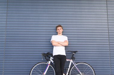 Genç adam bir beyaz T-shirt ile bir bisiklet duvar arka plan üzerinde duruyor ve kameraya görünüyor. Bir otoban scooter ile genç bir bisikletçi portresi.