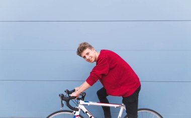 Mavi duvar bir arka plan üzerinde beyaz altıncı bisikletle oturan ve kameraya bakarak gülümseyen genç öğrenci. Mavi bir arka plan üzerinde mutlu öğrenci.