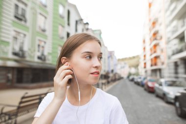 Yakın çekim portre müzik dinleyen bir gülümseyen kız gülümsüyor ve şehrin arka sokak doğru görünüyor. Şehirde yürüyüş ve kulaklık müzik dinlerken mutlu, şık kadın.
