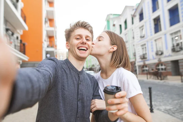一对年轻情侣的肖像在镇上的街道上自拍 微笑的年轻人用一杯咖啡在他的手采取自拍 他的女朋友亲吻在他的面颊 — 图库照片
