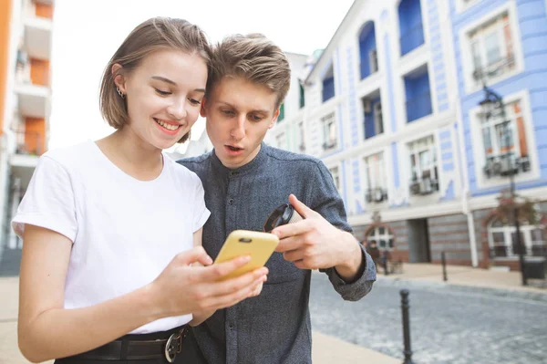 美丽的年轻夫妇使用智能手机在城市的街道上 女孩看着智能手机和微笑 年轻人显示手指 两个游客正在看智能手机上的路线 — 图库照片