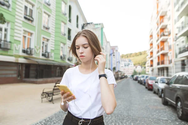 町の路上に立っている彼女の手でスマート フォンを見て ヘッドフォンで音楽を聴く深刻なスタイリッシュな女の子の肖像画 — ストック写真