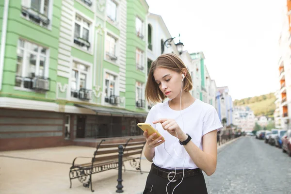 シャツに集中して魅力的な女の子はスマート フォンを使用し 古い街の背景でヘッドフォンで音楽を聴いています 流行に敏感な女の子 街歩き — ストック写真