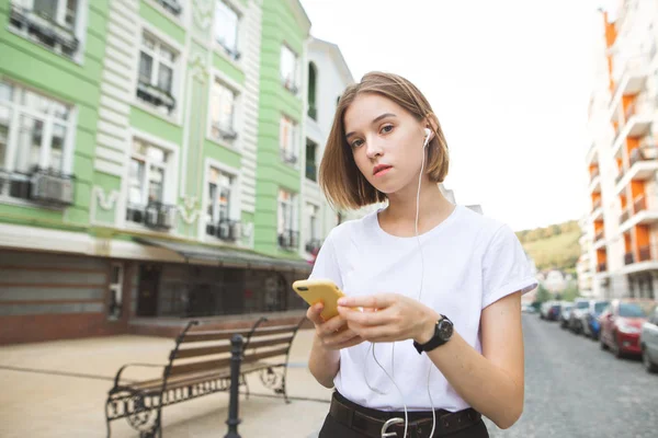 肖像画彼の手と通りの旧市街の背景にヘッドフォンでスマート フォンの側に立って カメラを探している魅力的な女の子 町散歩に白い シャツを着た女の子ヒップスター — ストック写真