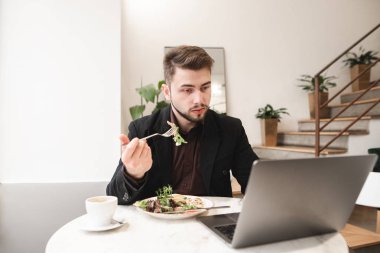 İş adamı kafede öğle yemeği için bir dizüstü bilgisayar kullanarak. Elbiseli meşgul adam bir restoranda yemek yiyen ve bilgi üstünde a laptop izler.