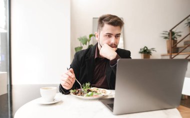 Bir tabak salata ve bir bardak kahve ile bir kafede bir masada oturan konsantre bir adam portresi ve bir dizüstü bilgisayar kullanarak. Meşgul bir adam bir restoranda bir dizüstü bilgisayar üzerinde çalışan öğle yemeği saat.