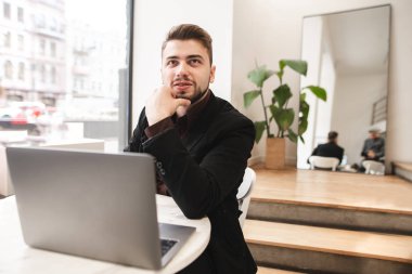 Gülümseyen adam bir takım ve rahat bir restoran bir tablo üzerinde bir dizüstü bilgisayar ile oturup bir sakal. Mutlu meşgul adam üstünde bir defter bir kafede çalışıyor.