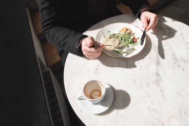 Bir fincan kahve ve bir tabak salata masanın üzerinde. Adam elinde çatal ve bıçak ile bir salata yiyor. Üstten görünüm. Sağlıklı gıda. Öğle yemeği molası. Işık ve gölge. Boşaltmak