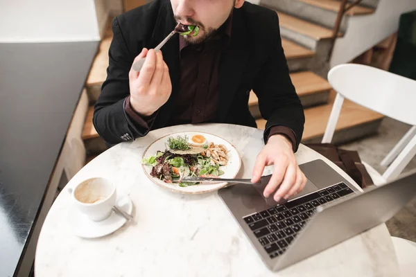Business man sitting with a laptop in a cafe and eating salad, a cup of coffee on the table. Busy man dishes in a cafe with healthy food and works on a laptop.