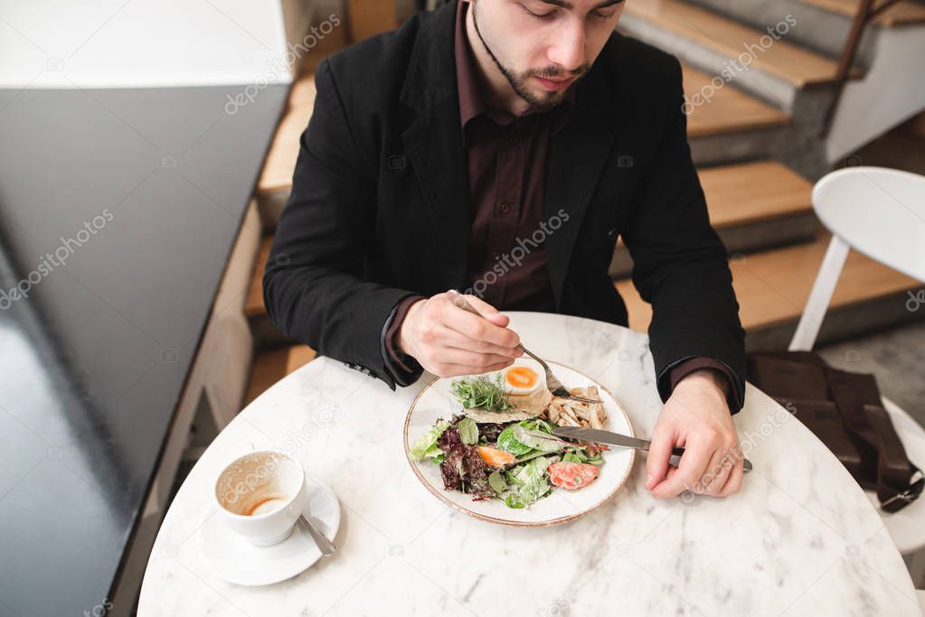 Business man sits in a cozy restaurant and eats a healthy meal, salad, a cup of coffee standing on the table. Top view. Man dishes with salad at a cafe at a break