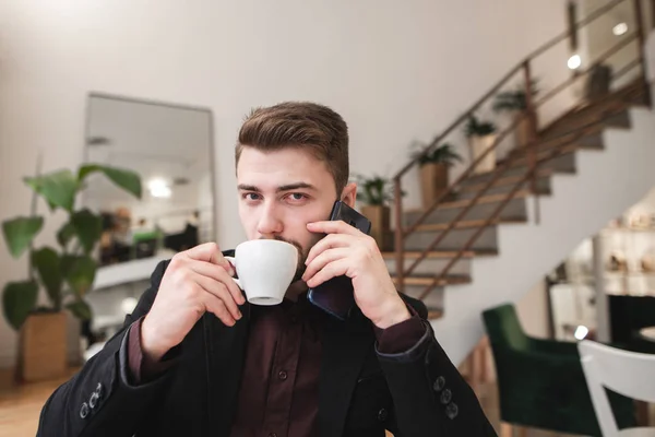Handsome man in a suit drinks coffee in a cozy restaurant, speaks by phone and looks at the camera. Portrait of a business man talking on the phone in a cozy cafe.