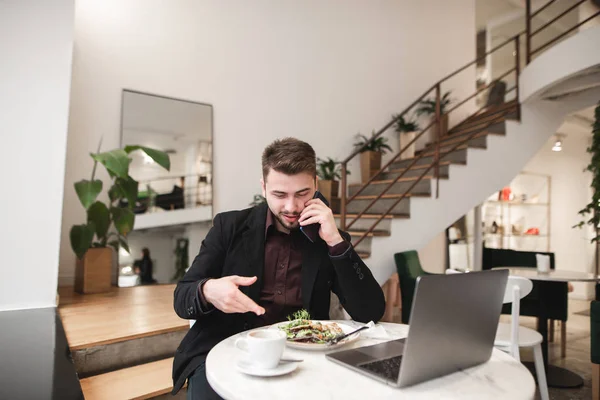 Positive man is sitting in a cafe with a laptop, coffee, food on the table, talking on the phone and smiling. Busy man works at a cafe for dinner. Handsome business man working in a cafe.