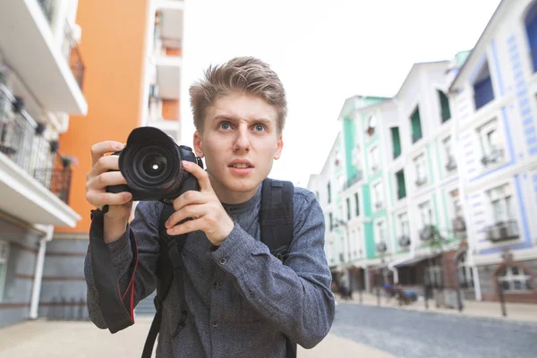 Street portrait of a funny young photographer with a camera in his hands against the backdrop of a beautiful town street. Young tourist in a shirt and with a camera looks seriously.