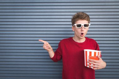 Kırmızı bir kazak ve patlamış mısır onun elinde bir fincan ile karanlık bir arka plan üzerinde duran ve onun parmak tarafında gösterilen beyaz 3d gözlük hayran genç adam. Sinema kavramı. Boşaltmak. Duvar