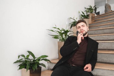 Bir takım elbise ve bitkiler yakınındaki merdivenlerde odada oturuyor ve smartphone üzerinde konuşurken bir sakal, ciddi bir adam portresi. Sol boşaltmak için