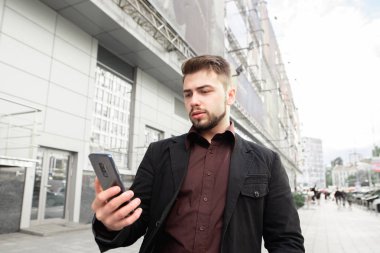 Portre sakal ve modern mimarinin arka sokakta duran ve bir smartphone kullanarak bir takım elbise ile bir iş adamı. Smartphone kavramı. Adam bir taksi çağırır.