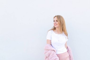 Çekici gülümseyen kız şık hafif giysiler ve kulaklık üzerinde beyaz bir arka plan teşkil etmektedir ve yana doğru boşaltmak üzerinde görünüyor. Beyaz bir duvar arka planı poz mutlu moda kız.