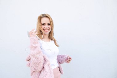 Bir genç şirin giysiler, kulaklık ile pozlar ve arka plan beyaz bir duvar Smartphone'da mutlu, trendy kız gülümser ve kamera görünüyor. Sevimli genç kadın. Boşaltmak