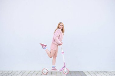 Mutlu kız pembe elbise, beyaz bir duvar, gülümsüyor arka plan üzerinde pembe tekme scooter üzerinde sürmek ve kameraya benziyor. Boşaltmak