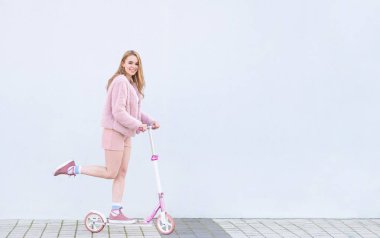 Mutlu, moda kız yayınlanan pembe elbise rides bir pembe tekme scooter üzerinde görünüyor içine belgili tanımlık fotoğraf makinesi ve gülümsüyor, beyaz bir arka plan üzerinde izole. Reklam için yer. Boşaltmak
