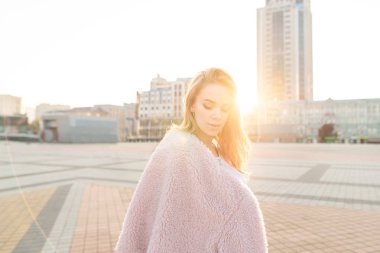 Şık bir model pembe elbiseleri günbatımı arka planda sokakta sokak portresi. Sevimli kız pembe elbiseli üzerinde akşam şehir manzarası arka kameraya poz veriyor.