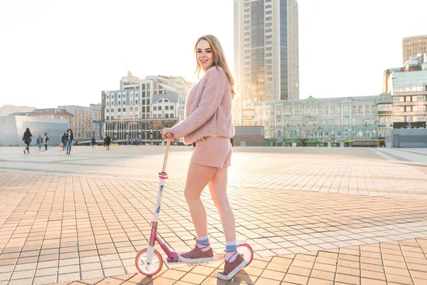 Girl in a pink fur coat and shorts stands with a kick scooter against the background of a city landscape at sunset, looks at the camera and smiles. Student girl goes on a kick scooter in the city.