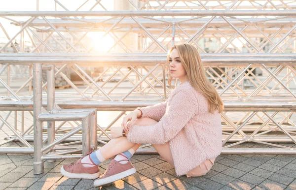 ピンク服夕日の背景に地面に座って よそ見スタイリッシュな魅力的な女の子 歩道に座っているピンクの毛皮のコートで魅力的な女の子 — ストック写真