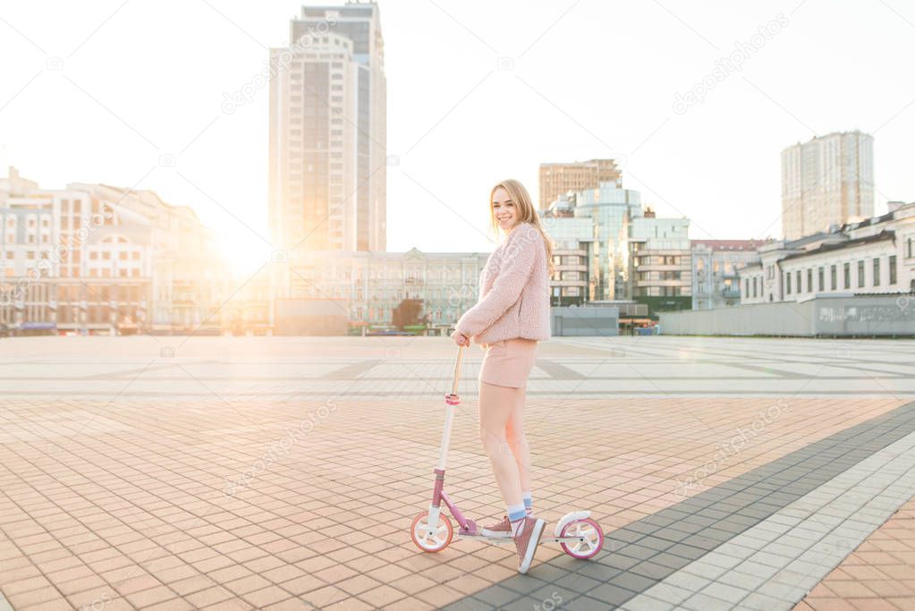 Attractive stylish blonde girl walking on a kick scooter in the city against the background of the sunset, looking into the camera and smiling. Evening riding on a scooter. Ecological transport.