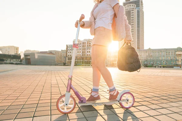 夕日の通りの背景手にバックパックでスクーターにピンクのかわいい十代の少女 女の脚 バックパックの彼の手とピンクのスクーター 太陽と都市の背景 — ストック写真