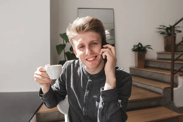 Student speaks by telephone and drinks coffee.Closeup portrait of a young attractive man dressed in a shirt sits in a cozy cafe, communicates on the phone, drinks coffee, look the camera and smiles