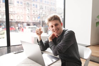 Bir dizüstü bilgisayar ve bir fincan kahve onun elinde hafif bir kafede otururken, kamera seyir ve gülümseyen gülümseyen genç bir adam portresi. Bir dizüstü bilgisayar ile bir masada oturan ve kafede kahve içme öğrenci