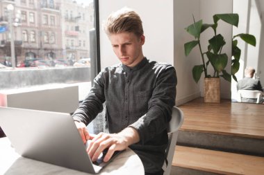 Bir gömlek konsantre genç adam rahat bir kafede oturur ve bir dizüstü bilgisayarda çalışır. Bir dizüstü bilgisayar ve bir kafede otururken, ekrana bakarak ve klavyede yazarak kahve