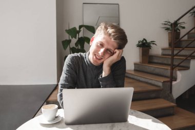 Bir dizüstü bilgisayar ve bir fincan kahve ile parlak bir samimi kafede otururken, kameraya bakıyor ve gülümseyerek mutlu bir genç adam portresi. Bir kafede bir dizüstü bilgisayar ile mutlu öğrenci