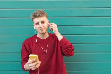 Gülümseyen güzel öğrenci parlak turkuaz duvar arka plan üzerinde elinde bir akıllı telefon ve kulaklık ile ayakta. Kulaklıklar müzik ve kamera arıyorum güzel bir adam portresi