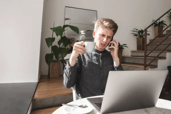 Serious young man speaks on a smartphone in a cozy cafe, drinks coffee and looks at the laptop screen. Concentrated young businessman works in a cafe.