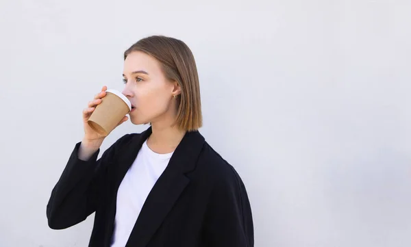 Attractive woman in a suit drinks coffee from a paper cup on a white background and looks to the side. Business woman drinks coffee. Copyspace