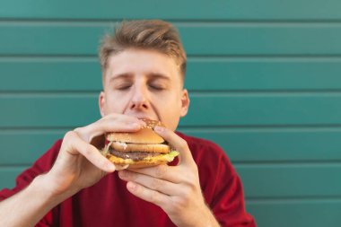 Turkuaz duvar arka planı kapalı gözlerle hamburger yeme genç bir adam portresi kapatın. Aç öğrenci renkli bir arka plan üzerine bir hamburger yiyor.