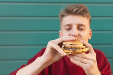 Turkuaz duvar arka planı kapalı gözlerle hamburger yeme genç bir adam portresi kapatın. Aç öğrenci renkli bir arka plan üzerine bir hamburger yiyor. Boşaltmak