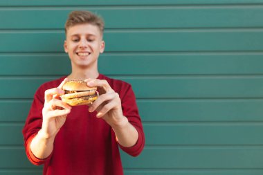 Genç adam gülümseyen bir renkli duvar arka planı elinde hamburger ile duruyor. Fast food. Genç Genç bir burger onun elinde. Burger üzerinde odaklanmak