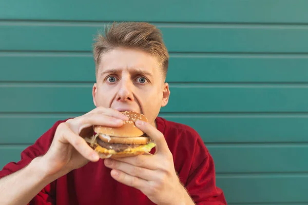 一个吃汉堡的年轻人的近照在绿松石墙的背景下看着镜头 饥饿的学生吃一个汉堡 快餐概念 — 图库照片