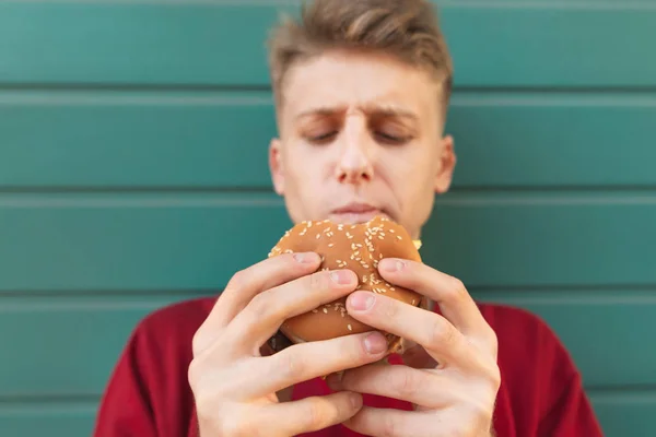 一个美丽的年轻人的肖像 手里拿着一个开胃的汉堡 背景是绿松石 把注意力集中在汉堡上年轻人在街上吃快餐 — 图库照片