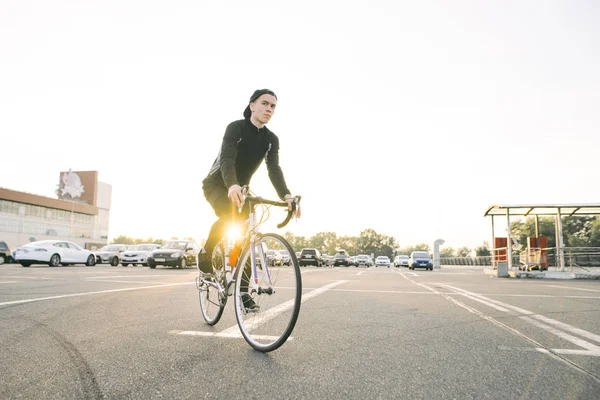 Genç adam koyu spor giyim ve bir kap beyaz bisiklet güneş park yerinde giyer. Bir bisikletçi öğrenci portresi. Bisiklete binme hobi.