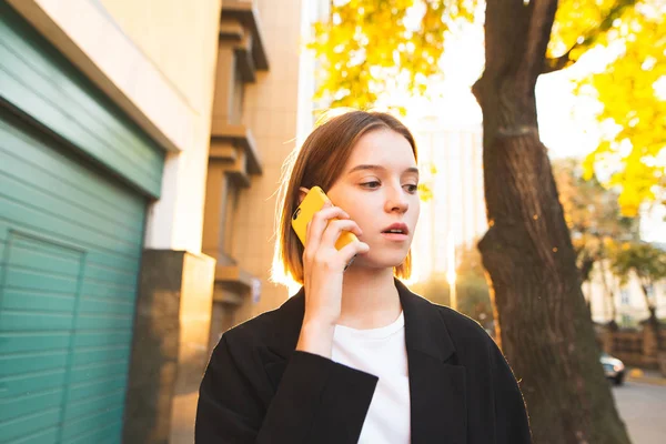 Serious attractive business woman in a suit calling on a smartphone on a bright city background. Outdoor portrait of a young business woman with backlight sunlight.