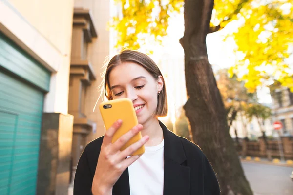 Street portrait of a happy young girl who uses a smartphone on the street. Smiling girl with a smartphone on the background of a sunny city landscape. Bright portrait.