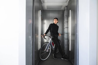 Spor giyim genç adam asansörde bir bisiklet ile standları ve aşağı iner.