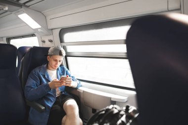 Concentrating young man goes by train and uses a smartphone. Tourist in a blue jacket and cap looks at the screen of a smartphone while on a train trip by the window. clipart