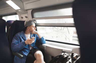 Student with a smartphone in his arms goes by train and looks in the window. Guy travels with a modern train and uses a smartphone. clipart