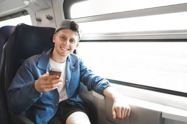 Mutlu genç traveler bir treni pencerenin yakınında oturan, bir smartphone kullanarak, kameraya bakıyor ve gülümsüyor. Genç adam tren, internette bir gezi trenler.