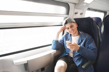 Portrait of a smiling boy in casual clothes sitting in a train near the window with a smartphone in his hands, looking into the camera and smiling. Happy train trainer smiles. clipart